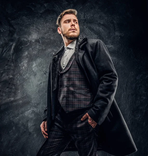 Un hombre elegante vestido con un elegante traje y abrigo posando con una mano en un bolsillo contra una pared de textura oscura —  Fotos de Stock