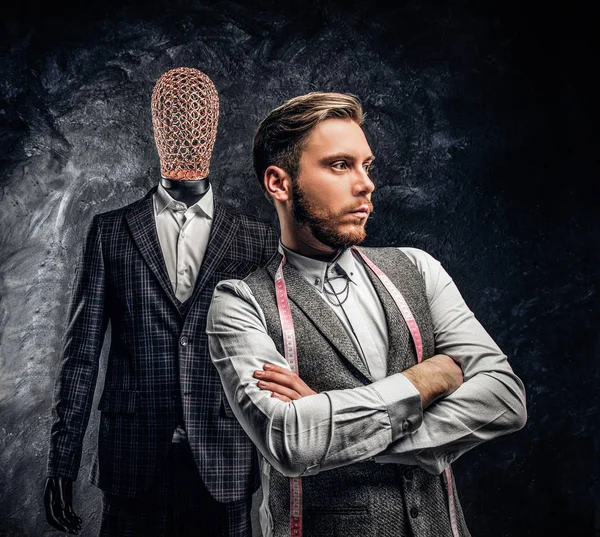 Um jovem designer de moda confiante posando ao lado de um manequim em terno exclusivo feito sob encomenda dos homens em um estúdio de alfaiate escuro — Fotografia de Stock