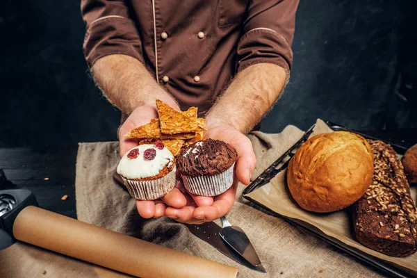 Manos masculinas sosteniendo deliciosas magdalenas junto a una mesa con panes y baguettes —  Fotos de Stock