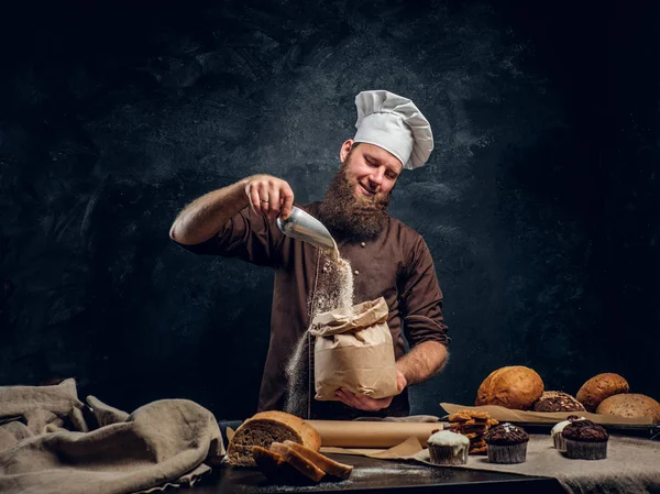 A bearded baker wearing a cook uniform sprinkling some flour in bag, standing next to a table, decorated with delicious bread loaves, baguettes — Stock Photo, Image