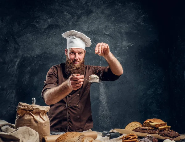 Wesoły Brodaty baker, posypując trochę mąki na jego świeże muffin obok tabeli — Zdjęcie stockowe