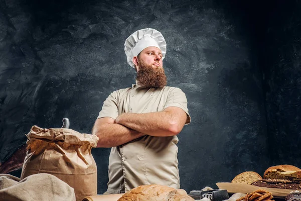 Um padeiro barbudo vestindo um uniforme de pé com os braços cruzados ao lado de uma mesa, decorado com deliciosos pães, baguetes — Fotografia de Stock