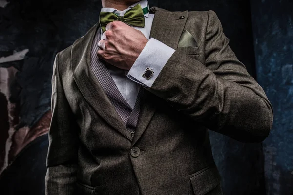 Un joven elegante vestido con un traje elegante corrige su pajarita contra una pared de textura oscura — Foto de Stock