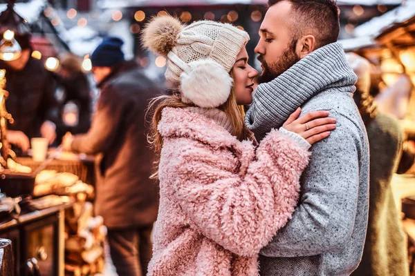 Casal apaixonado, um casal elegante vestindo roupas quentes abraçando juntos e olhando uns aos outros na feira de inverno em um momento de Natal . — Fotografia de Stock