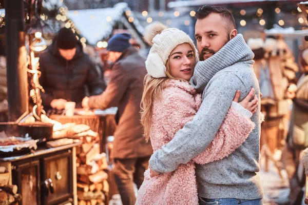 Una encantadora pareja enamorada, abrazándose juntos y mirando una cámara mientras están de pie en la feria de Navidad — Foto de Stock