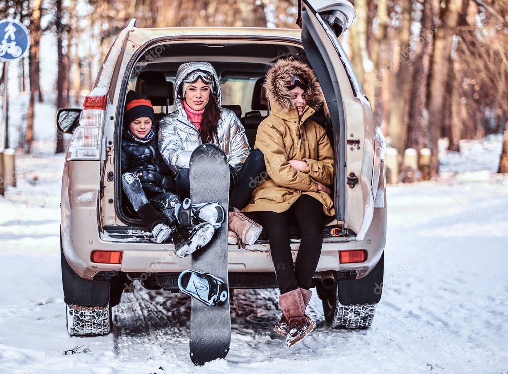 A beautiful woman and her sons dressed in warm clothes sitting on the trunk of a car