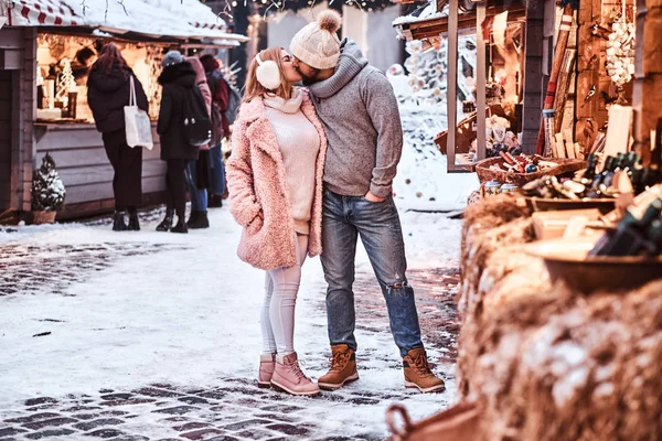 Um casal atraente apaixonado vestindo roupas quentes elegantes de mãos dadas e beijando enquanto estava na feira de inverno em um momento de Natal . — Fotografia de Stock