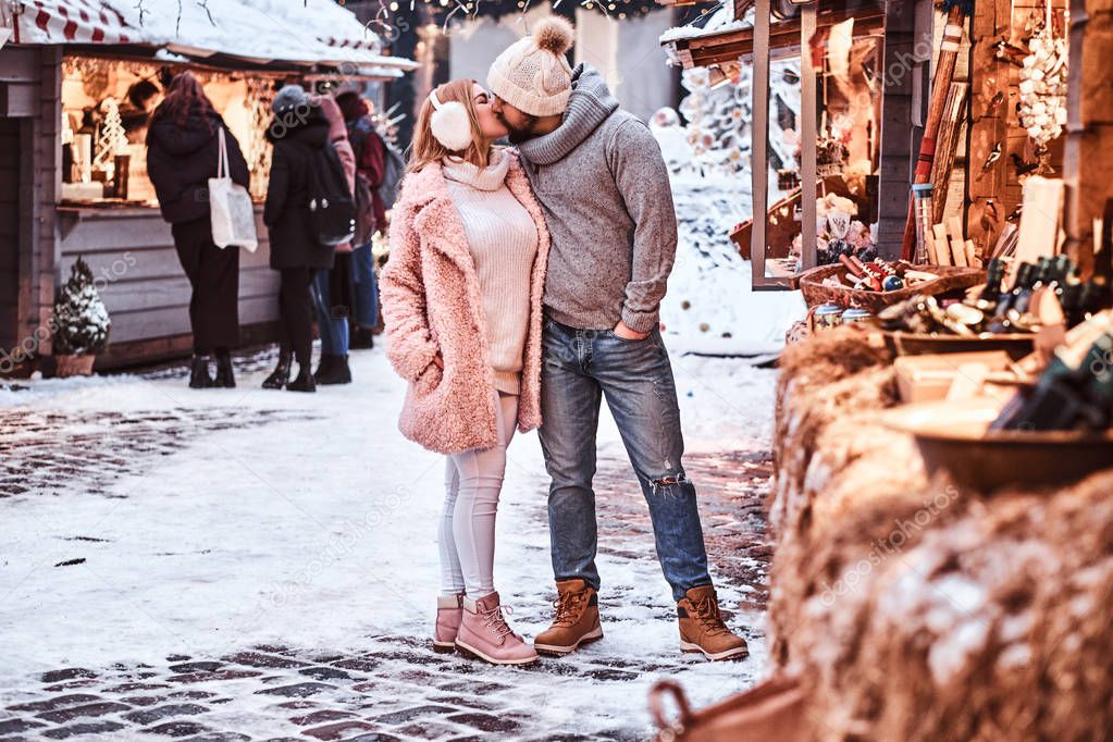 An attractive couple in love wearing stylish warm clothes holding hands and kissing while standing at the winter fair at a Christmas time.