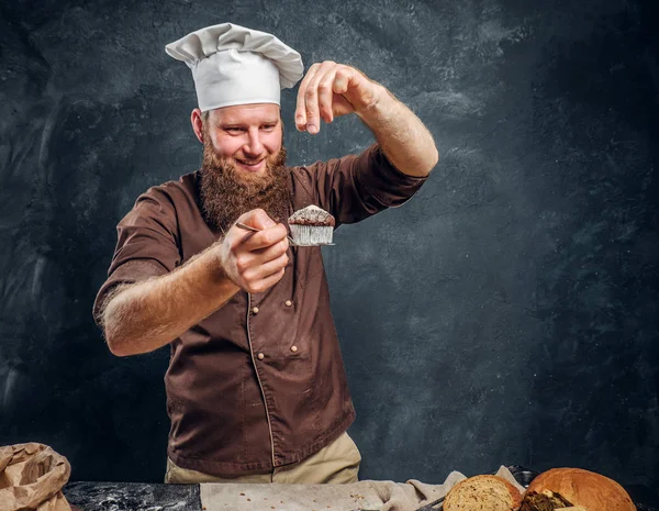 Alegre padeiro barbudo polvilhando um pouco de farinha em seu queque acabado de fazer ao lado de uma mesa — Fotografia de Stock