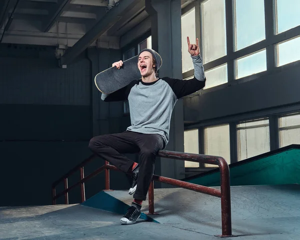 Fröhlicher Mann mit Hemd und Hut, der sein Brett hält, sitzt auf einer Schleifschiene im Skatepark drinnen. — Stockfoto