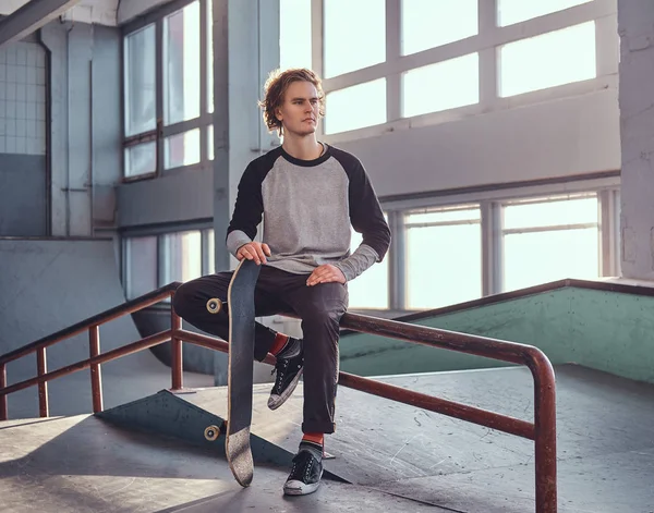 Young red-haired skateboarder holding his board while sitting on a grind rail in skatepark indoors. — Stock Photo, Image