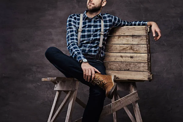Imagen recortada. Hombre pasado de moda con una camisa a cuadros con tirantes sentados en un andamio de madera — Foto de Stock