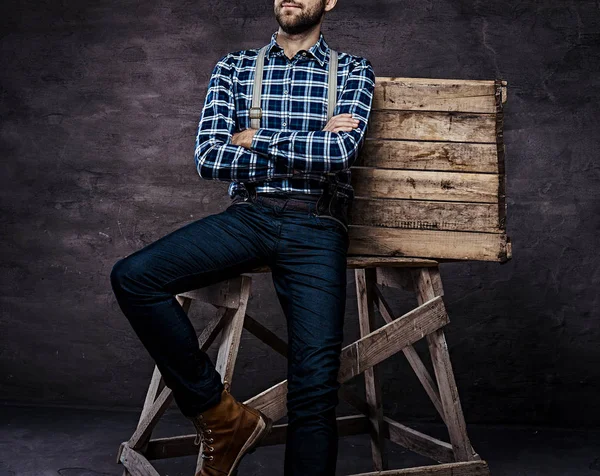 Imagen recortada. Hombre pasado de moda con una camisa a cuadros con tirantes con los brazos cruzados sentado en un andamio de madera — Foto de Stock