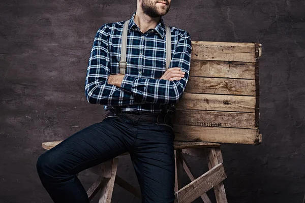 Imagen recortada. Hombre pasado de moda con una camisa a cuadros con tirantes con los brazos cruzados sentado en un andamio de madera — Foto de Stock