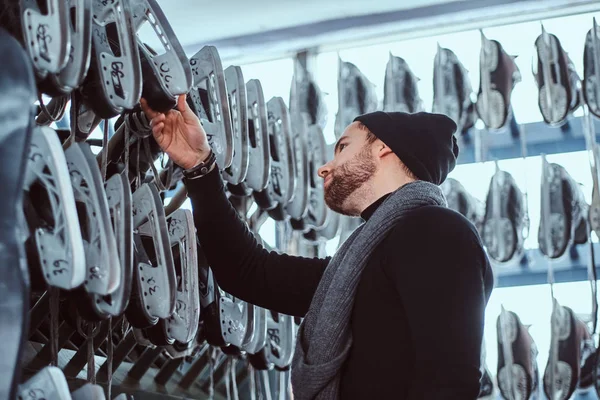 Ein junger Mann in warmer Kleidung wählt ein Paar Schlittschuhe in einem Skate-Verleih im Inneren. — Stockfoto