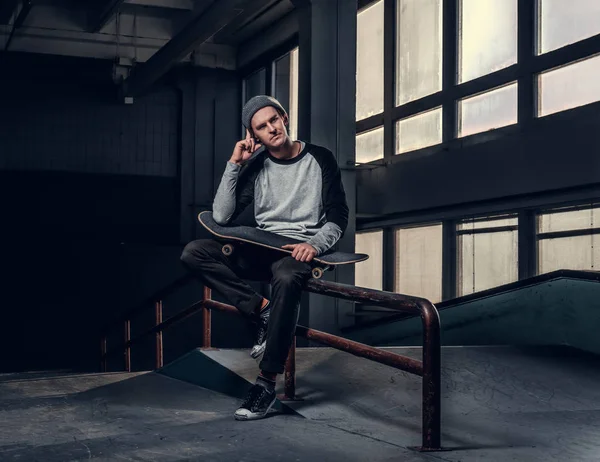 Jonge knappe man dragen shirt en hoed houden van zijn bestuur zittend op een spoor van de sleur in skatepark binnenshuis — Stockfoto