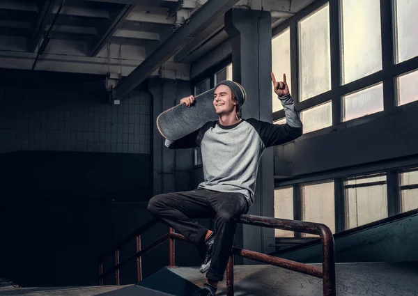 Vrolijke man dragen een shirt en een hoed zijn bestuur, zittend op een spoor van de sleur in skatepark binnenshuis te houden. — Stockfoto