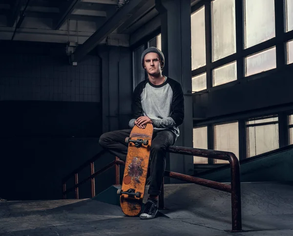 Jonge knappe man dragen shirt en hoed houden van zijn bestuur zittend op een spoor van de sleur in skatepark binnenshuis — Stockfoto