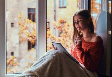 Girl in a warm sweater covered her legs with a blanket, makes notes in her notebook sitting on the window sill next to the open window clipart