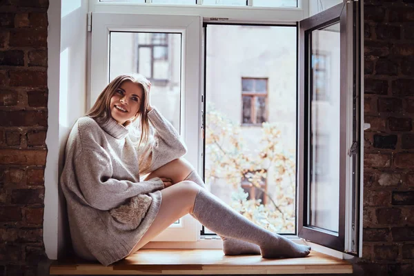 Cute girl in a warm sweater and socks sits on the window sill next to the window open
