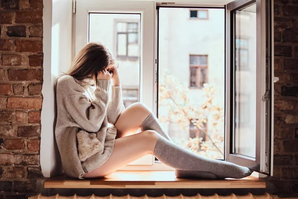 Милая девушка в тёплом свитере и носках сидит на подоконнике рядом с открытым окном — стоковое фото