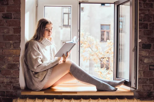 Linda chica en un suéter caliente y calcetines lee un libro sentado en el alféizar de la ventana al lado de la ventana abierta — Foto de Stock