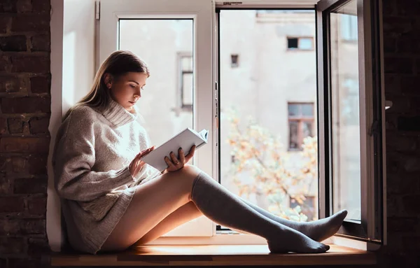 Aranyos lány egy meleg pulóver és zokni elolvassa a könyvet, az ablak párkányára nyitott ablak mellett ülve — Stock Fotó