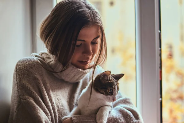 Pretty girl in a warm sweater hugs her favorite cat sitting on the window sill next to the open window