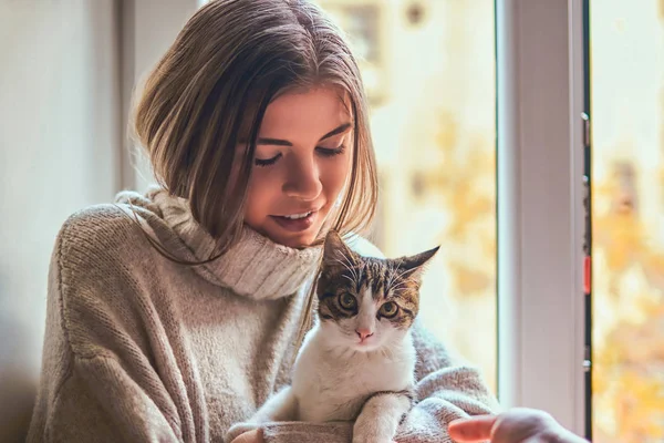 Красива дівчина в теплому светрі обіймає свого улюбленого кота, що сидить на підвіконні біля відкритого вікна — стокове фото