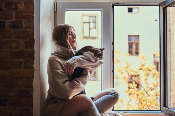 Pretty girl in a warm sweater hugs her favorite cat sitting on the window sill next to the open window