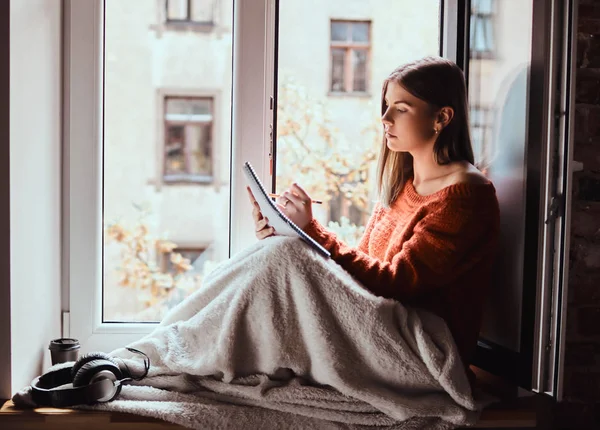 暖かいセーターの女の子が毛布で彼女の足をカバー、ノートは、開いているウィンドウの横にある窓の下枠の上に座って彼女のノート — ストック写真