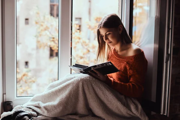 Mladá žena v teplém svetru zahrnuty nohy s dekou, při pohledu na foto album, sedí na okenním parapetu — Stock fotografie