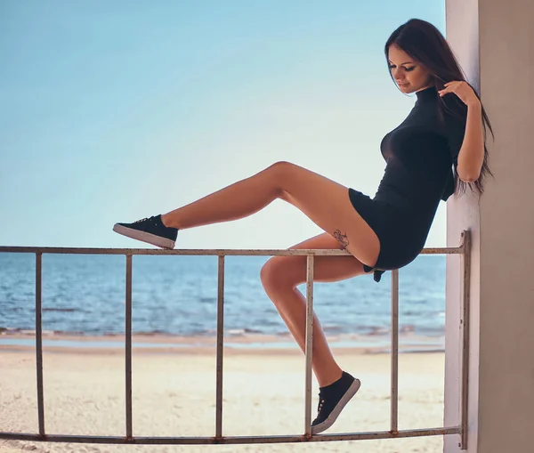 Krásná dívka s tetováním na noze sedět na zábradlí proti krásné moře, pobřeží — Stock fotografie