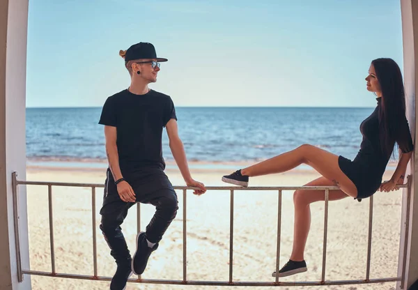 Casal amoroso em roupas elegantes sentar no corrimão e olhar uns para os outros contra a bela costa do mar — Fotografia de Stock