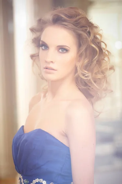 Porträt einer charmanten Frau mit stylischer Lockenfrisur, die in einem blauen Kleid neben einem Spiegel posiert. Schönheit, Mode, Mode. — Stockfoto