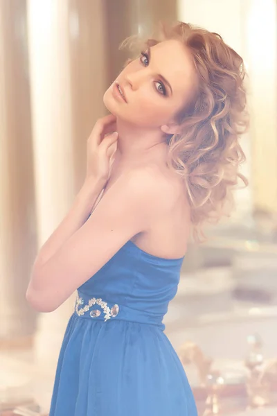 鏡の隣にポーズ青いドレスを着てスタイリッシュな巻き毛のヘアスタイルの魅力的な女性の肖像画。美容、流行、ファッション. — ストック写真