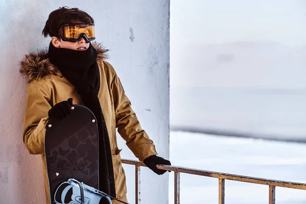 Un jeune snowboardeur vêtu de vêtements chauds et de lunettes de protection assis sur une rampe près de la plage enneigée — Photo
