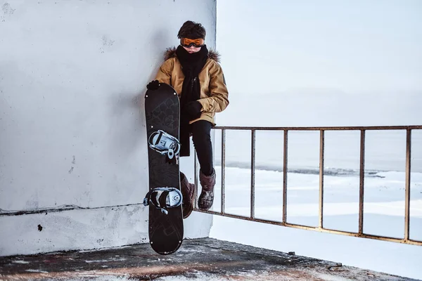 Een jonge snowboarder gekleed in warme kleding en veiligheidsbril zittend op de leuning in de buurt van het besneeuwde strand — Stockfoto