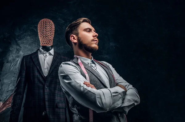 Un joven diseñador de moda confiado posando junto a un maniquí en traje exclusivo hecho a medida para hombre en un estudio de sastre oscuro — Foto de Stock