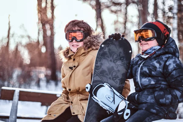 Due fratelli adolescenti vestiti con abiti caldi seduti su una panchina con uno snowboard in un parco — Foto Stock
