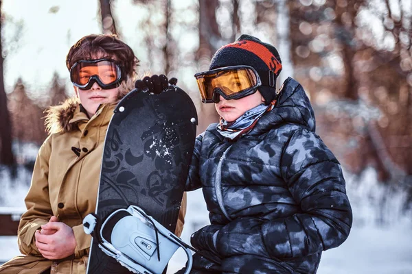 Due fratelli adolescenti vestiti con abiti caldi seduti su una panchina con uno snowboard in un parco — Foto Stock