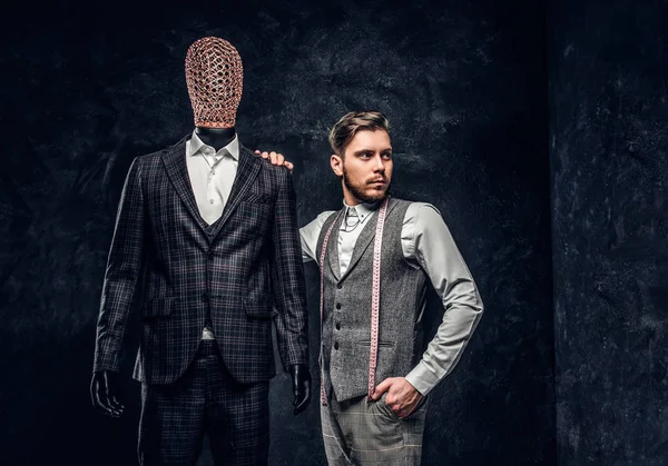 Un joven diseñador de moda confiado posando junto a un maniquí en traje exclusivo hecho a medida para hombre en un estudio de sastre oscuro — Foto de Stock