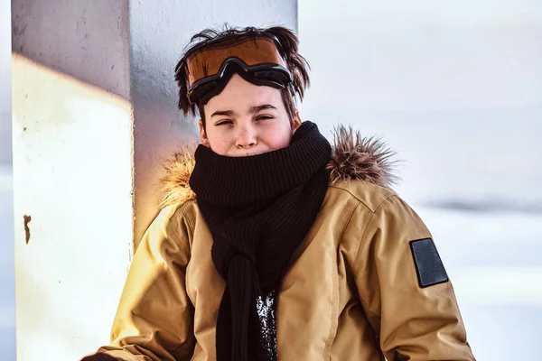 Un adolescent snowboarder vêtu d'une combinaison de neige et de lunettes de protection assis sur une rampe près de la plage enneigée — Photo