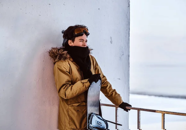 Un joven snowboarder vestido con ropa de abrigo y gafas protectoras sentado en la barandilla cerca de la playa nevada —  Fotos de Stock