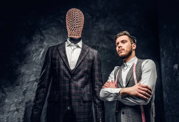 Een zeker jonge modeontwerper poseren naast een mannequin in exclusieve aangepaste gemaakte mens pak in een donkere kleermaker studio — Stockfoto