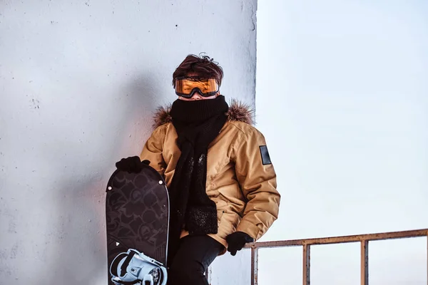 A young snowboarder dressed in warm clothes and protective goggles sitting on railing near the snowy beach — Stock Photo, Image