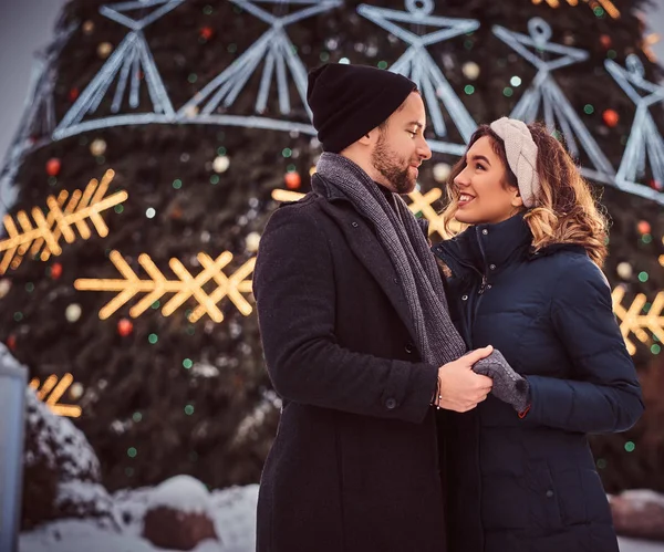 Pareja feliz vistiendo ropa de abrigo tomados de la mano y mirándose, de pie cerca de un árbol de Navidad de la ciudad, disfrutando pasar tiempo juntos . — Foto de Stock