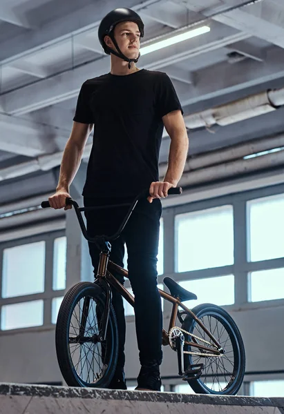 Retrato de un joven en casco protector con su bicicleta de pie en un parque de skate en el interior — Foto de Stock