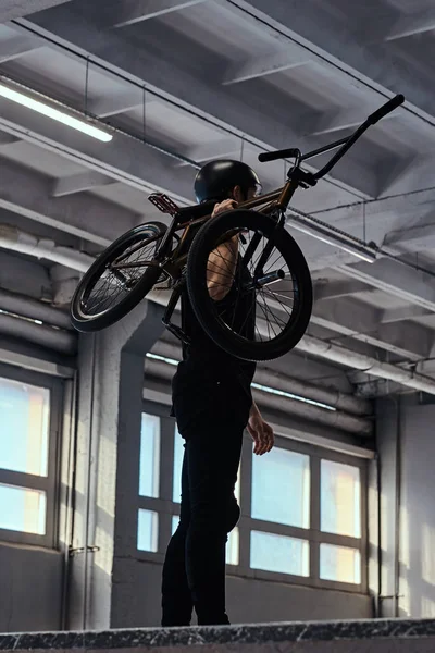 Jinete profesional de BMX en un casco de protección sosteniendo su bicicleta y mirando hacia otro lado en un parque de skate en el interior. Foto vertical — Foto de Stock