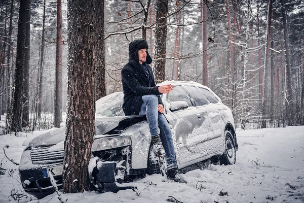 Samochód wpadł w poślizg i uderzył w drzewo na zaśnieżonej drodze. Kierowca siedzi na masce silnika i pali papierosa w oczekiwaniu Laweta — Zdjęcie stockowe